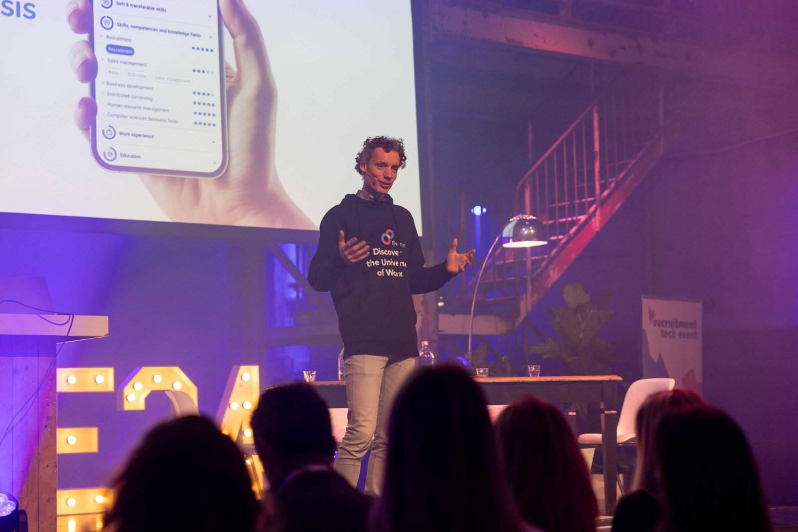 Laurens Waling in een donkere 8vance-hoodie op het paars verlichte podium van het Recruitment Tech Event, voor een scherm met een skillsprofiel op een telefoon