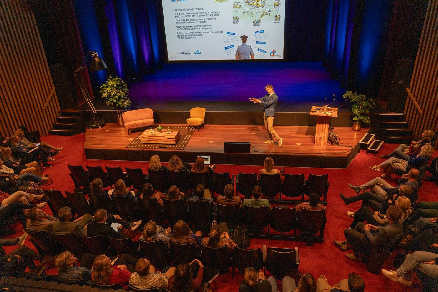 Zicht vanaf het balkon op een vol theater, met Laurens Waling alleen op het podium naast twee oranje fauteuils en een slide met arbeidsmarktcijfers